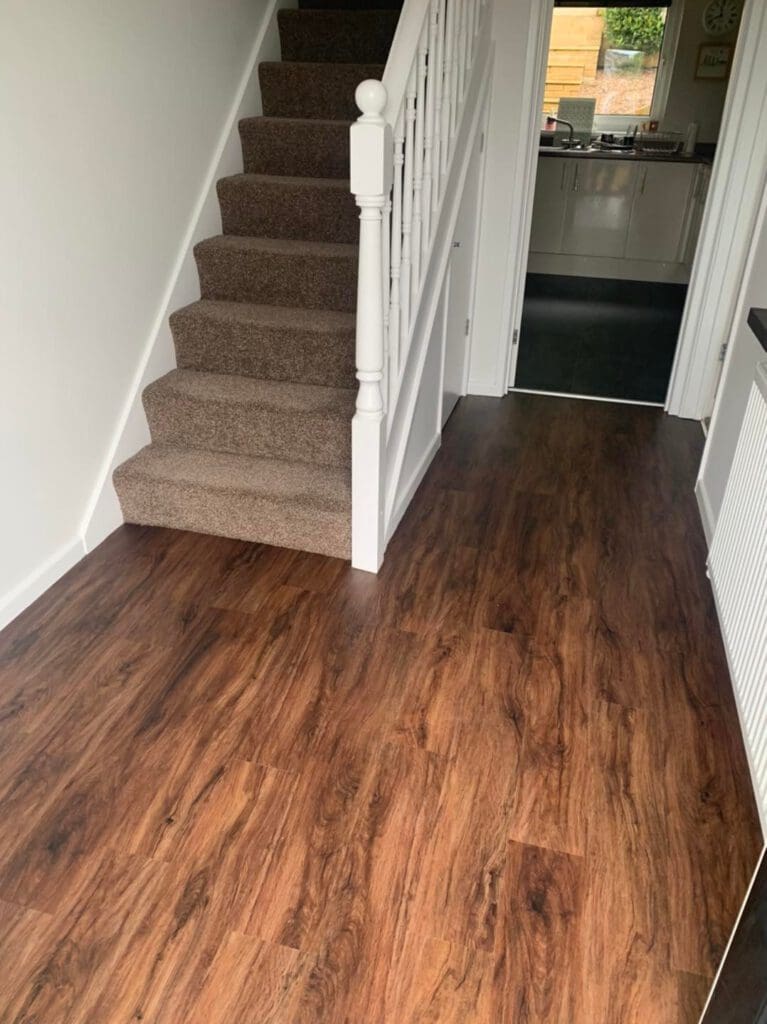 A hallway with dark wood flooring leads to a carpeted staircase on the left and a kitchen visible through a doorway at the end. White walls and a radiator are on the right side of the hallway.