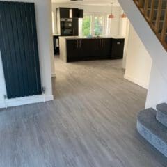 A modern interior featuring a staircase with grey carpeted steps, leading to an open kitchen with black cabinets, white countertops, and pendant lights. The floor has a grey wood appearance, and a tall dark radiator is mounted on the wall.