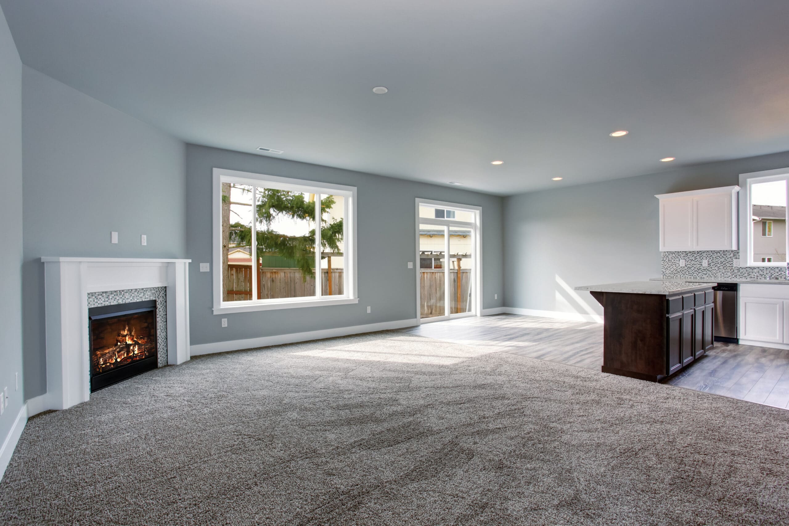 Spacious living room with carpeted floors, light gray walls, and a white fireplace. Large windows and glass doors let in natural light. The open floor plan leads to a kitchen with white cabinets and a dark island.