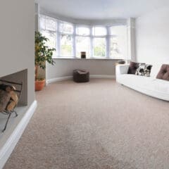 A bright, minimalist living room with a large bay window, white sofa with brown cushions, floor lamp, and potted plant. A beige carpet covers the floor, and a small pile of firewood is visible in a nook on the side.