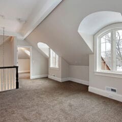 A spacious attic room with beige walls and brown carpet. It features large arched windows that allow natural light to fill the space. A staircase with a black and white railing leads down to the lower level.