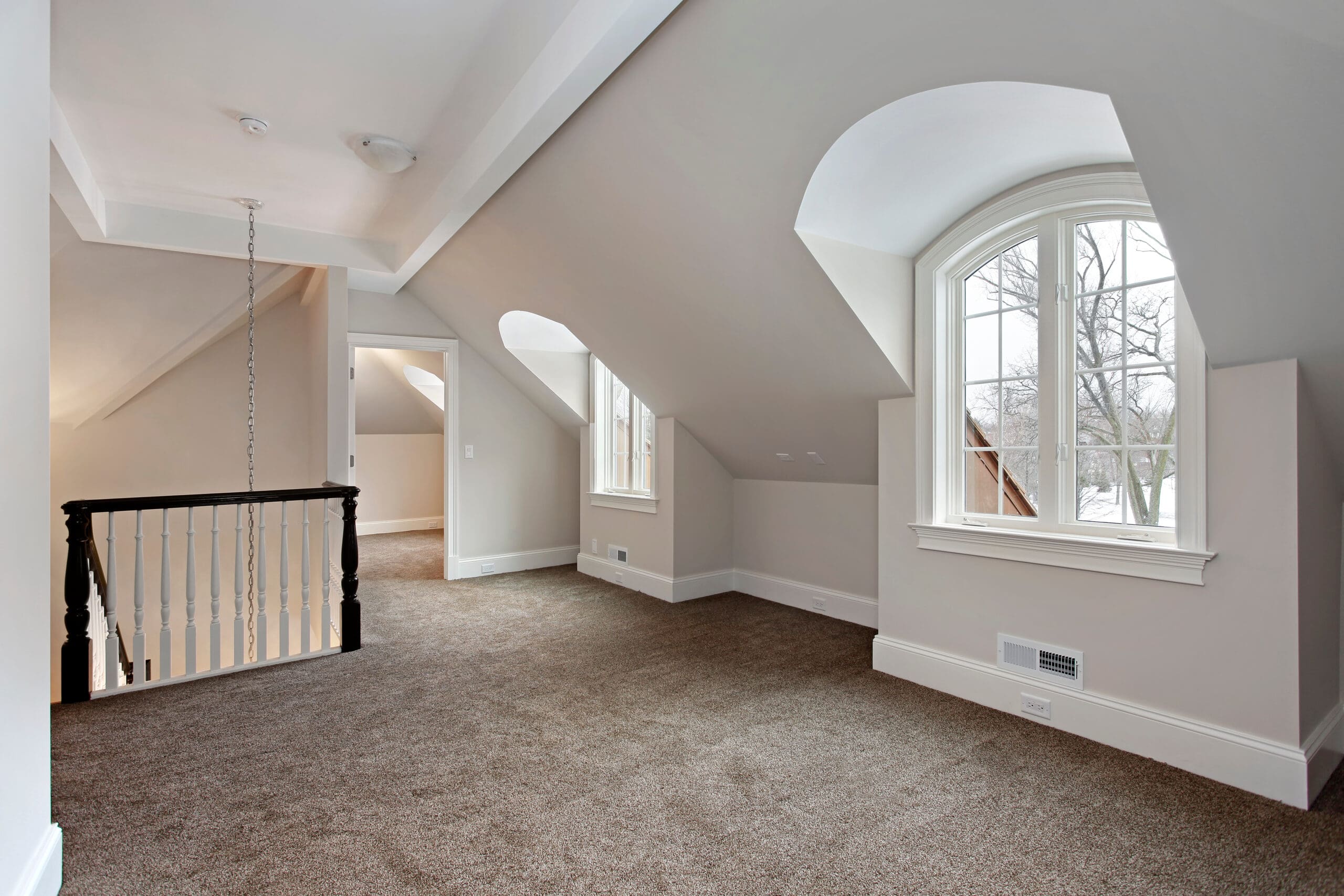 A spacious attic room with beige walls and brown carpet. It features large arched windows that allow natural light to fill the space. A staircase with a black and white railing leads down to the lower level.