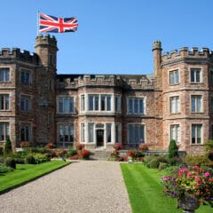 A historic, castle-like mansion with turrets and a large Union Jack flag on top. It is surrounded by landscaped gardens with colorful flowers and a gravel path leading to the entrance under a clear blue sky.