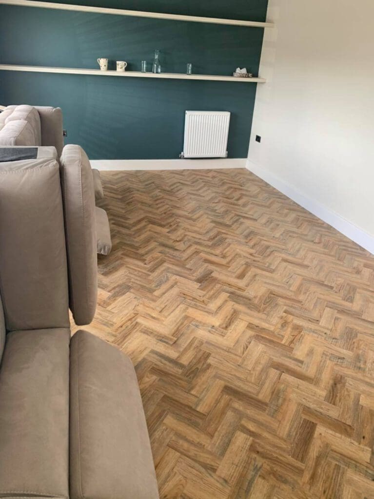A room with a herringbone wood floor, green wall with white shelves, and a beige sofa. Shelves display cups and vases, and a small white radiator is mounted beneath them.