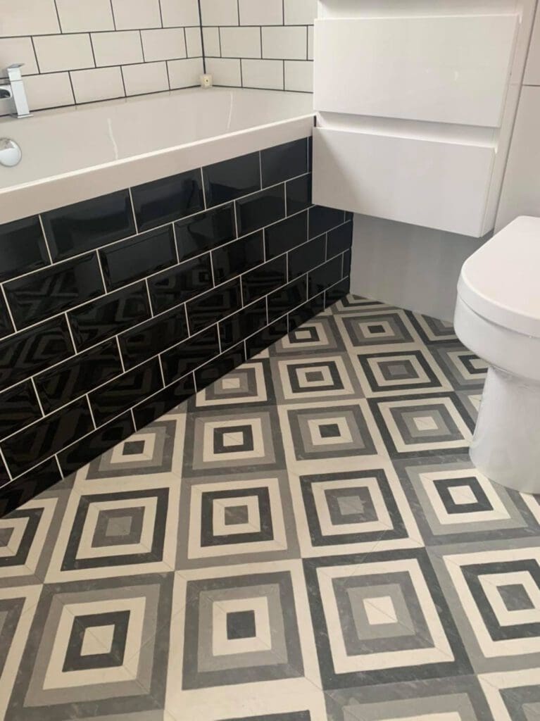 Bathroom with a modern design featuring a bathtub with glossy black subway tiles on the side and a geometric patterned floor in black, white, and gray. A white cabinet and toilet are also visible.
