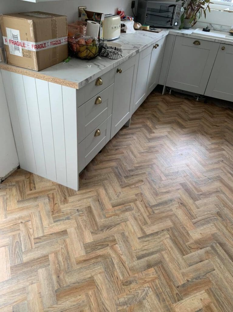 A kitchen with light gray cabinets and brass handles, featuring a box labeled "Fragile" on the counter. The floor displays a herringbone wood pattern. Appliances and a fruit bowl are also visible on the countertop.