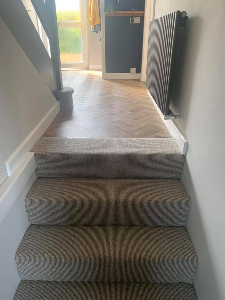 A staircase with grey carpet leads to a hallway with herringbone-patterned flooring. A dark vertical radiator is on the right wall. A door at the end of the hallway shows a glimpse of outdoor greenery.