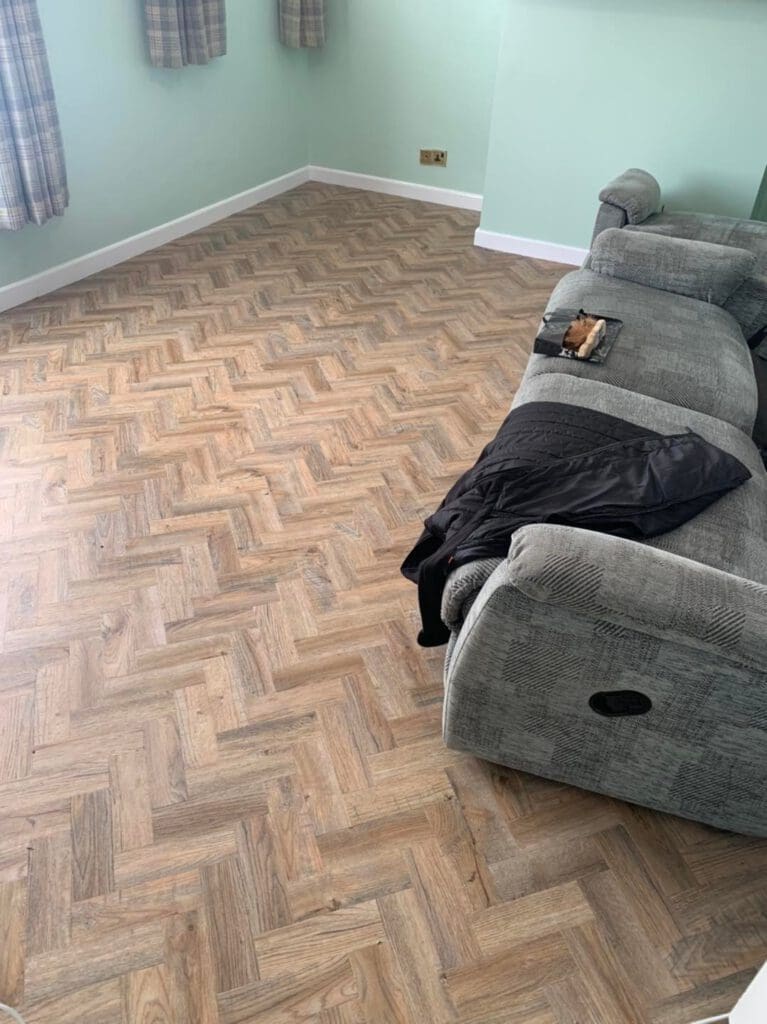 A room with light green walls featuring a chevron-patterned wooden floor. A grey couch with a black jacket draped over one arm is positioned in the corner. A small book or notebook is placed on the couch. The curtains are plaid design.