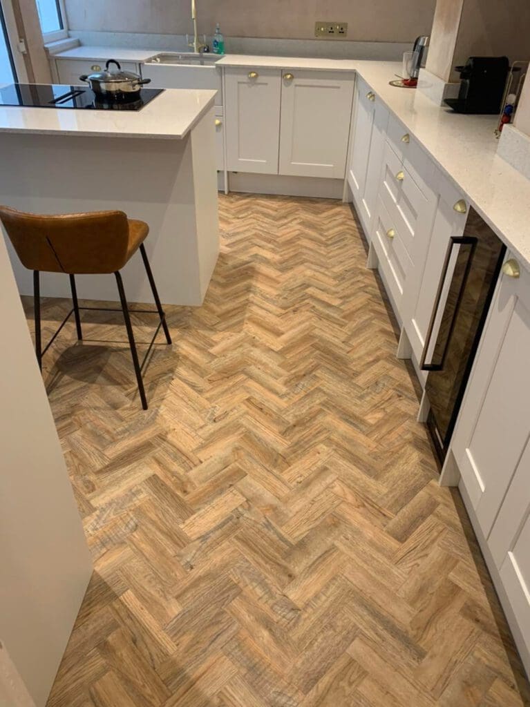 Modern kitchen with white cabinets, a wooden herringbone-patterned floor, and a central island with a brown bar stool. There is a stove on the island and an oven and small appliances along the counter. Bright and neatly arranged.