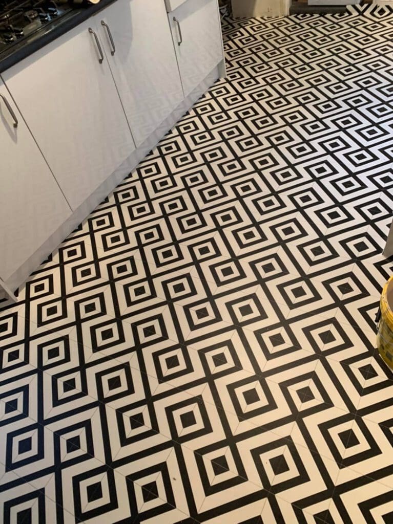 Kitchen floor with a bold black-and-white geometric pattern, featuring interlocking squares and diamonds. White cabinets are visible on the left side, adding contrast to the intricate tile design. A yellow object is partially visible on the right.