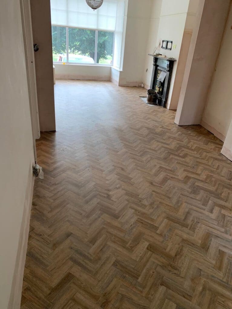 A room with light wood herringbone flooring, white walls, and large windows allowing natural light. A decorative fireplace with a mirror above it is on the right. The room appears unfurnished.