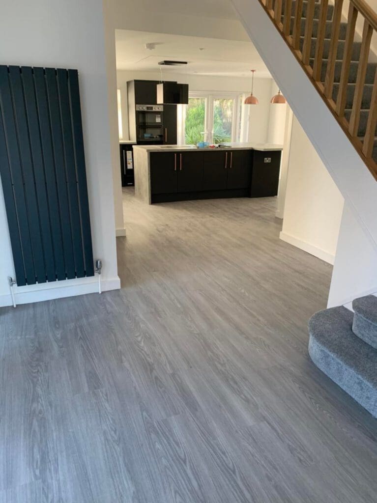 Open-plan interior with modern kitchen in the background. Features dark cabinets, light countertops, and pendant lights. Foreground shows gray wood flooring, a staircase with carpeted steps, and a vertical black radiator on the wall.