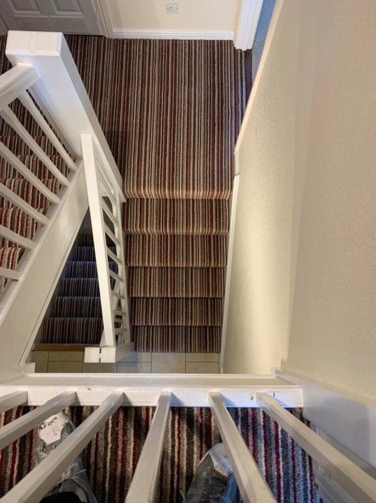 View from the top of a narrow staircase with white railings and brown striped carpet. The stairs lead down through a small landing and turn at a right angle. The surrounding walls are cream-colored.