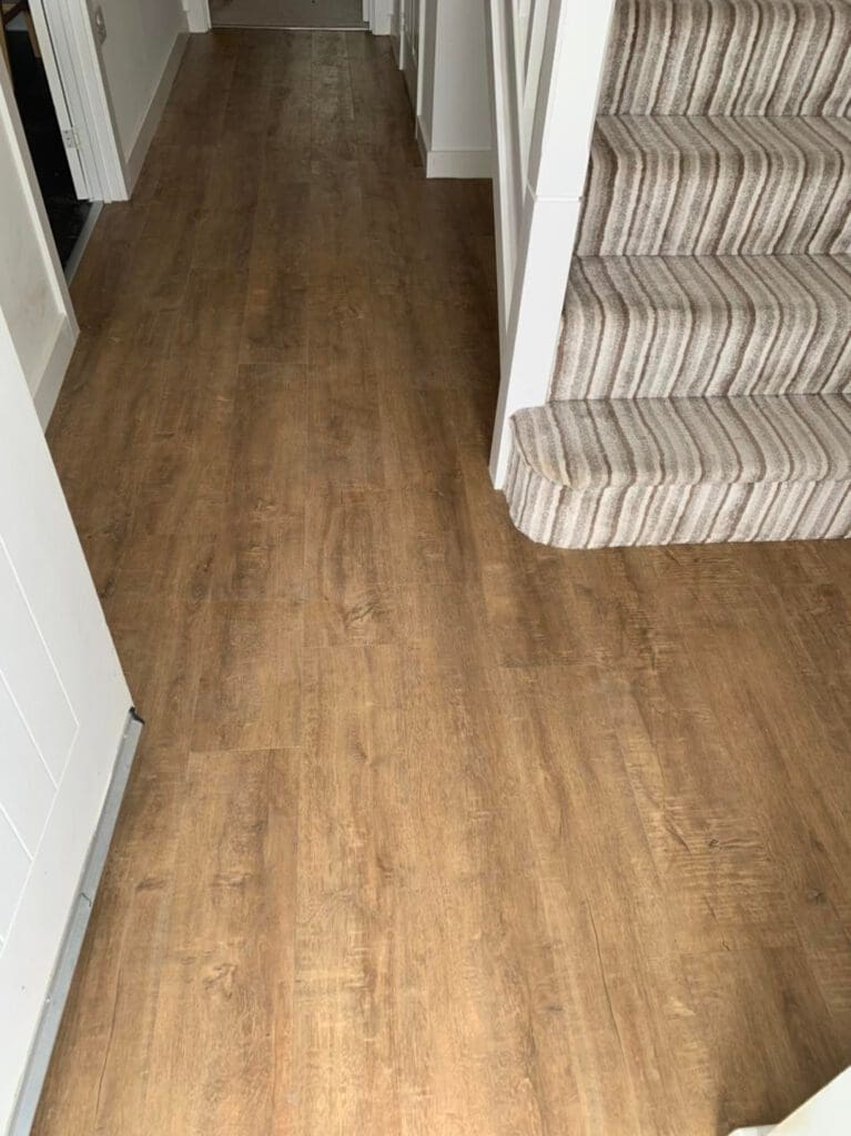 A hallway with wood-patterned vinyl flooring and a staircase covered in striped carpet. The space is bright and well-lit with white walls, and a portion of a door is visible at the end of the hall.