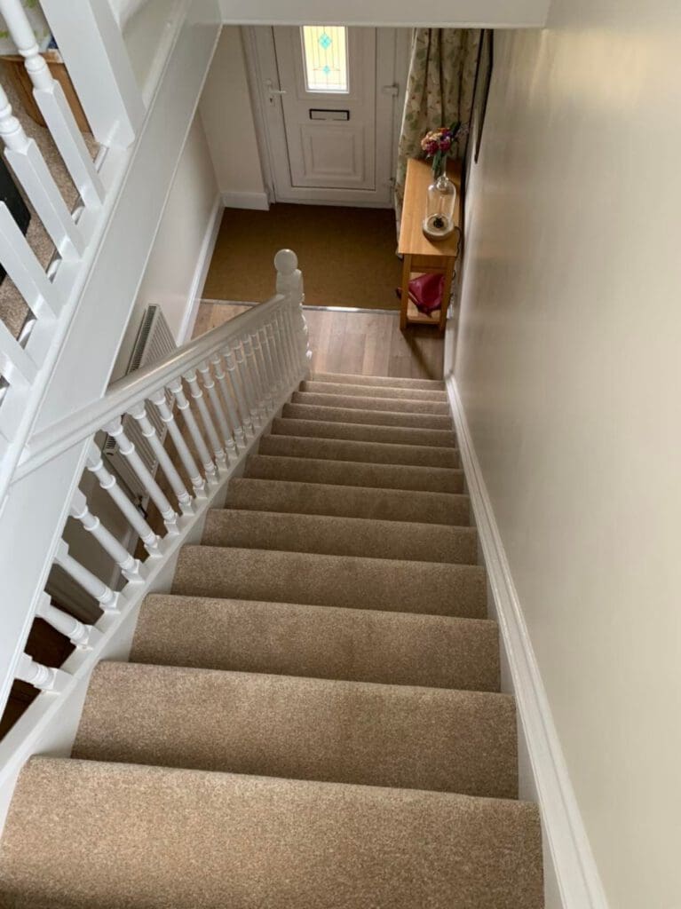 View from the top of a carpeted staircase leading down to a small foyer area. The foyer has a wooden table with a vase of flowers on it, and a white front door with a stained glass window panel is visible at the bottom.