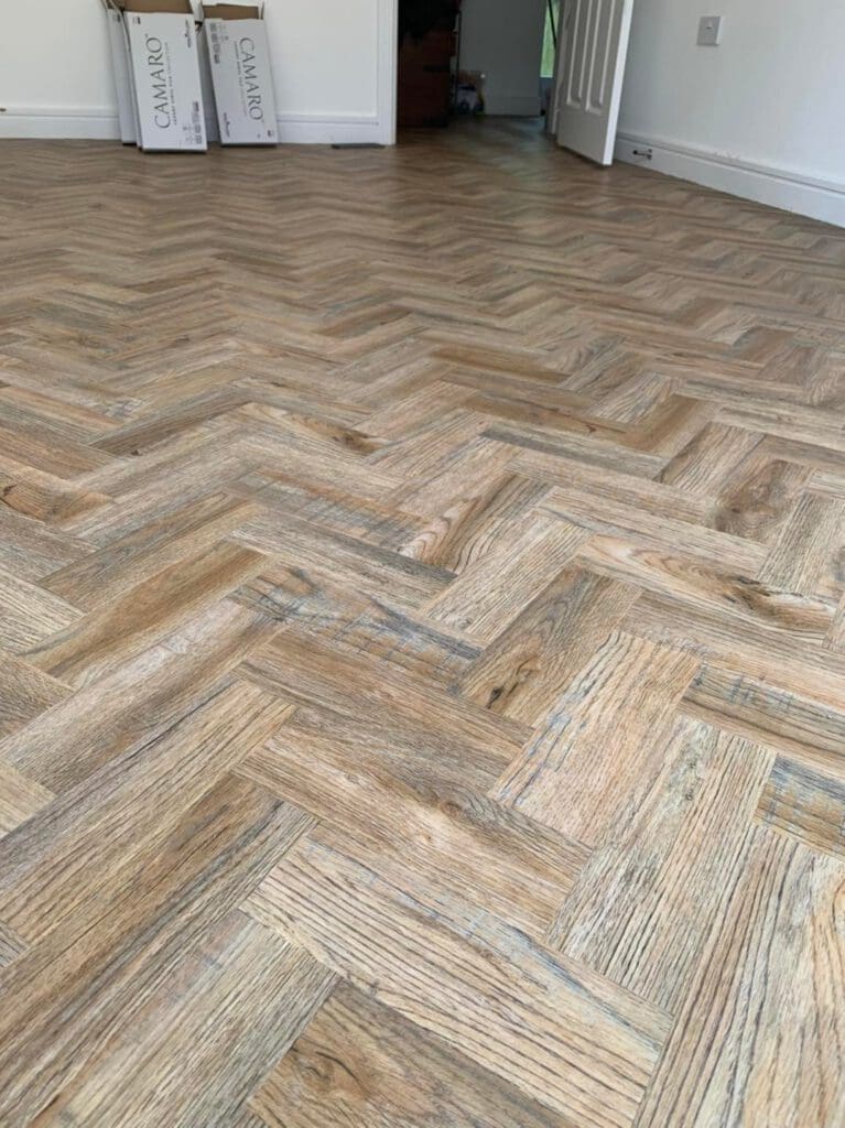 A room with a newly installed herringbone pattern wooden floor. The light wood tones add warmth to the space. In the background, two boxes labeled "Camaro" are stacked against the wall near an open door.