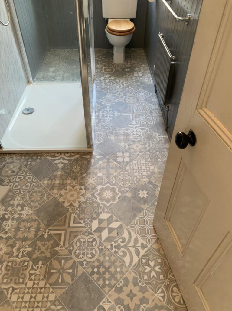 Bathroom with patterned gray and white tile floor, a glass shower stall on the left, and a toilet with a wooden seat at the far end. The walls are gray with vertical paneling, and a door with dark handles is partially visible.