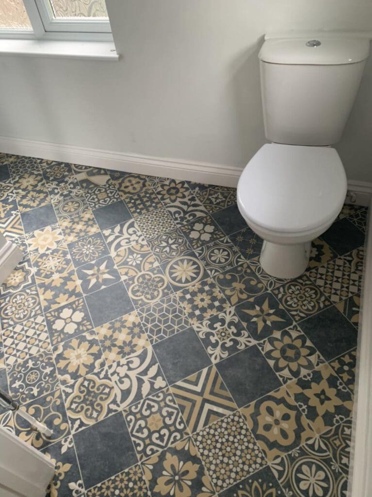 A bathroom with a white toilet next to a window. The floor is covered with blue and beige patterned tiles featuring various geometric and floral designs.