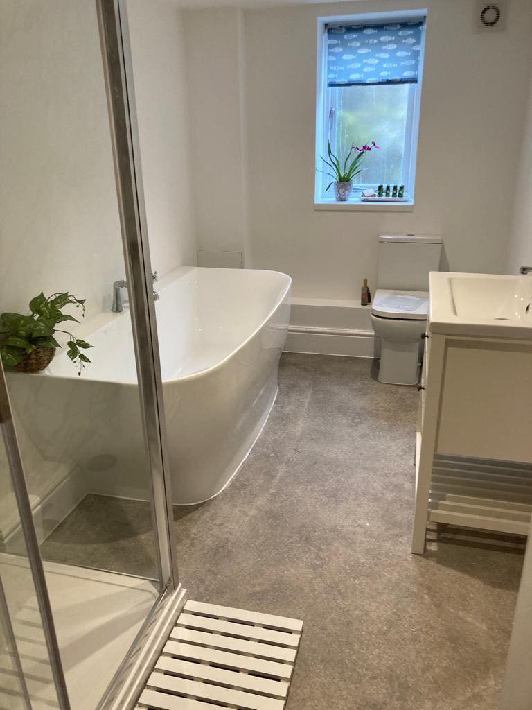 A modern bathroom with a white freestanding bathtub, a glass-enclosed shower, a toilet, and a sink with a vanity. There's a potted plant by the bathtub and a window with a patterned blind, bringing in natural light.
