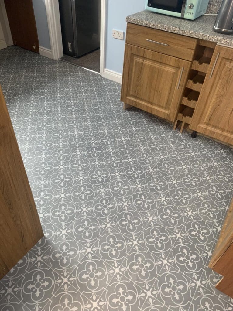Kitchen with wooden cabinets and patterned tile flooring featuring white floral designs on a gray background. A wine rack is partially visible, and a blue wall is in the background.