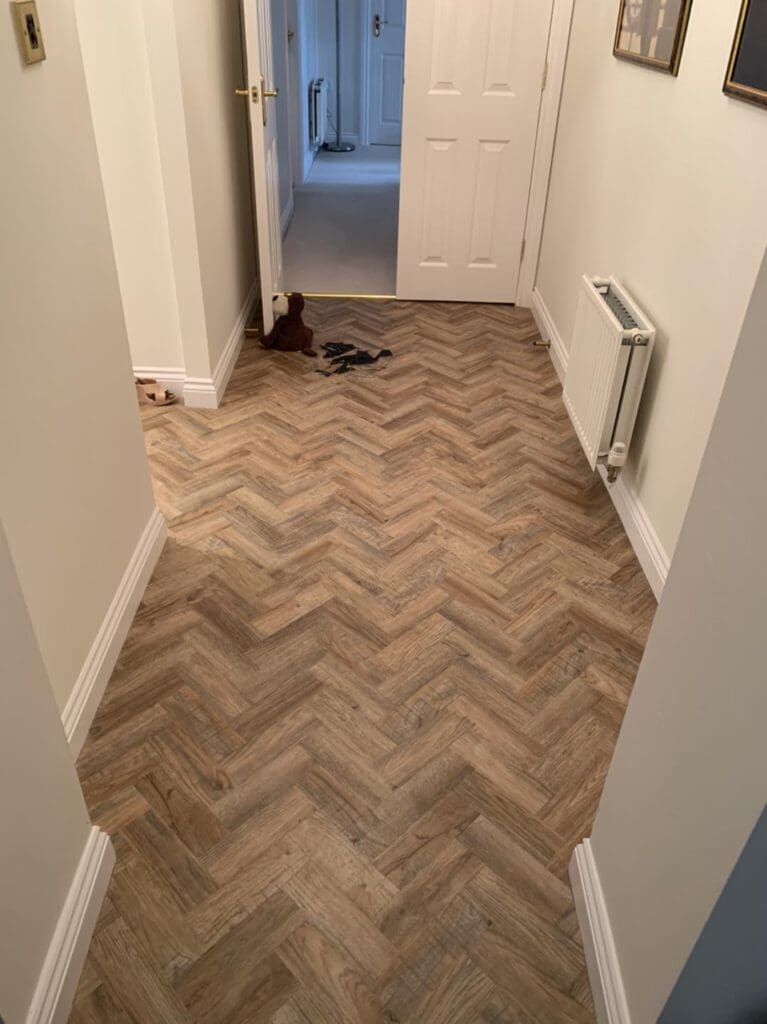 A hallway with herringbone-patterned wooden flooring. A white radiator is mounted on the right wall. The walls are adorned with framed pictures, and there's an open door at the end of the corridor. Some items rest on the floor near the door.
