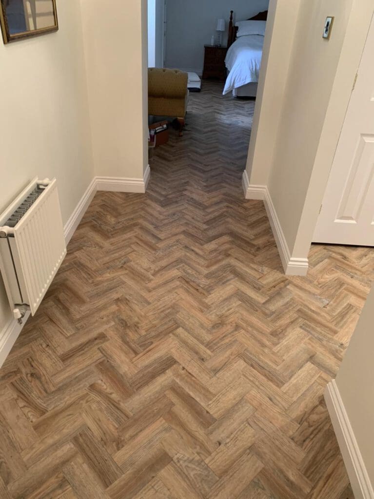 A hallway with wood-patterned herringbone flooring leads to a bedroom. White walls and a radiator are on the left, while an armchair is partially visible at the end near a bed with white linens.