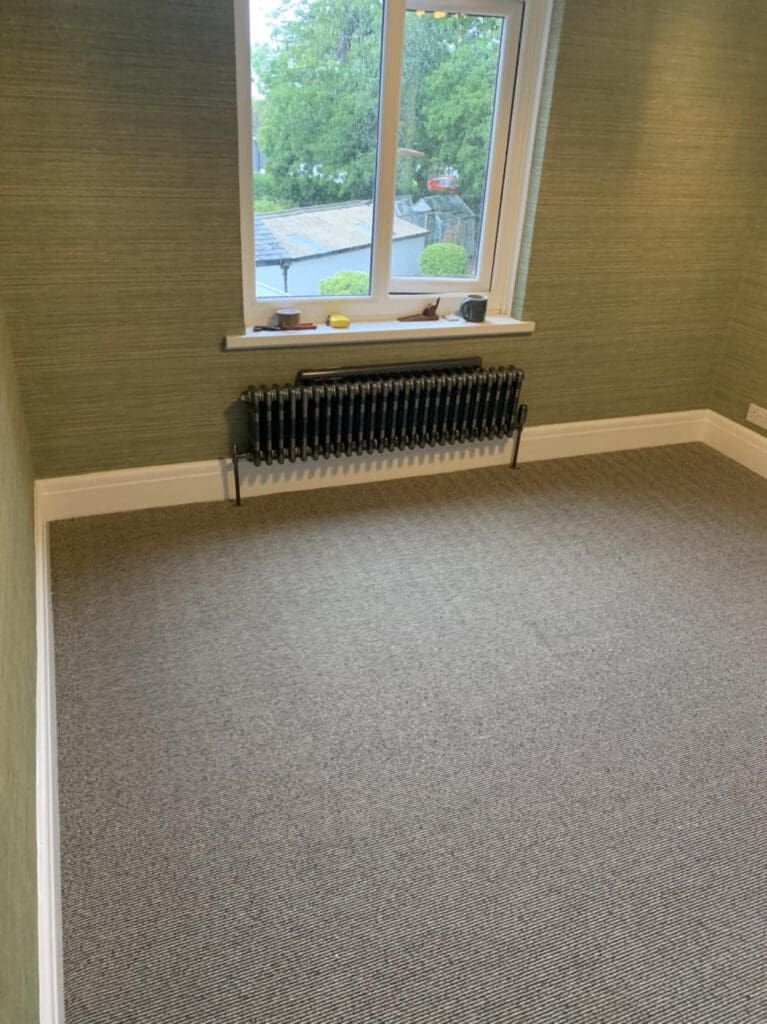 A small, empty room with green walls and a gray carpet. There is a window with a view of trees and rooftops, and a black radiator beneath it.