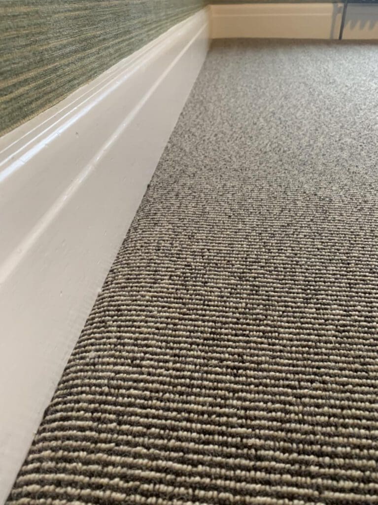 Close-up of a textured gray carpet meeting a white baseboard against a green wall. The perspective creates a diagonal line from foreground to background, highlighting the contrast between the carpet texture and the smoothness of the baseboard.